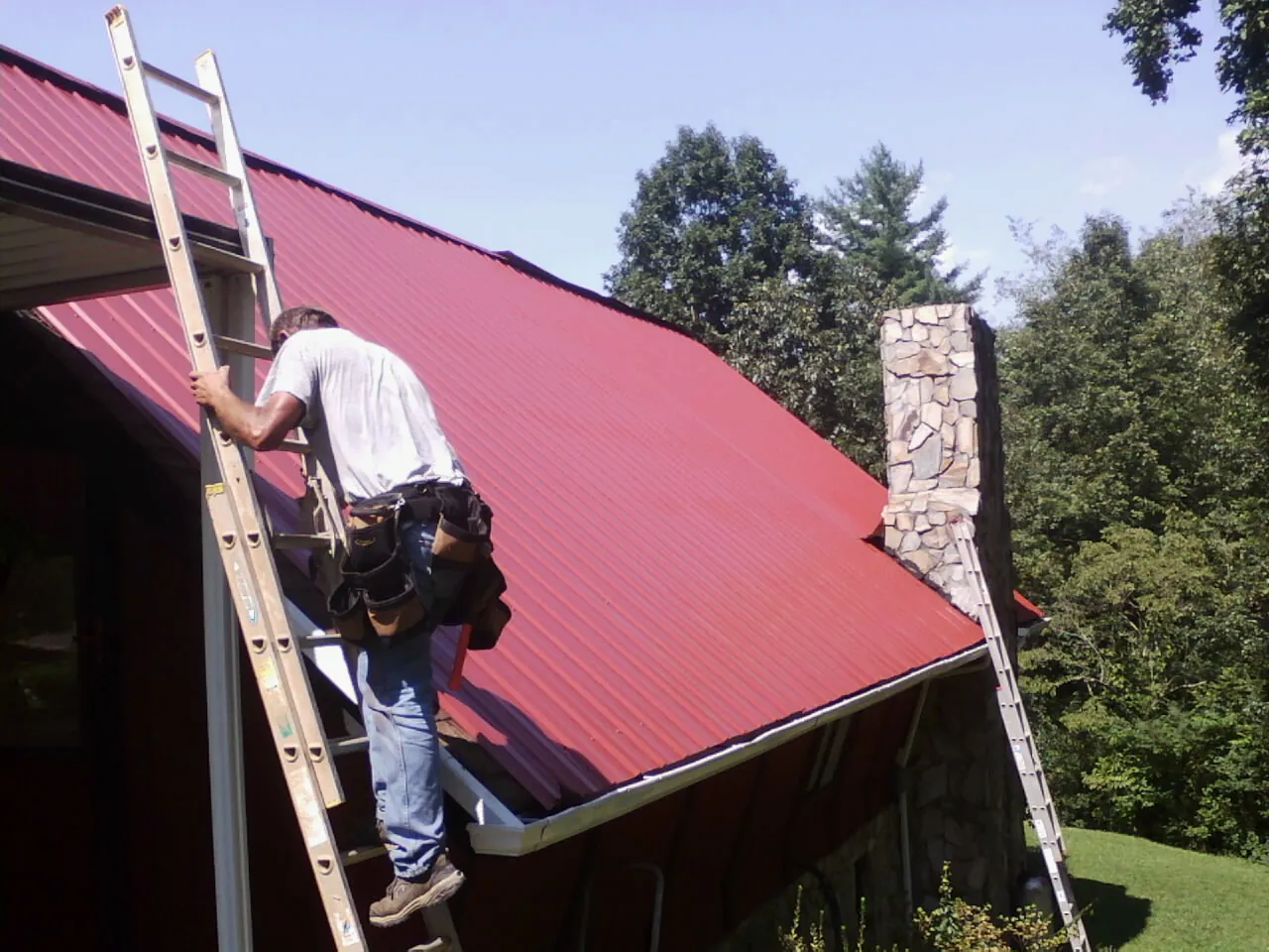 Roofer on a ladder beside a red metal roof and stone chimney — Soffit & Fascia Repair by Roofing Brothers in Snowmass Village